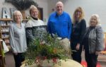 Five adults standing together indoors, smiling behind a round table with a green floral arrangement; casual setting with neutral walls and decorative shelf in the background.