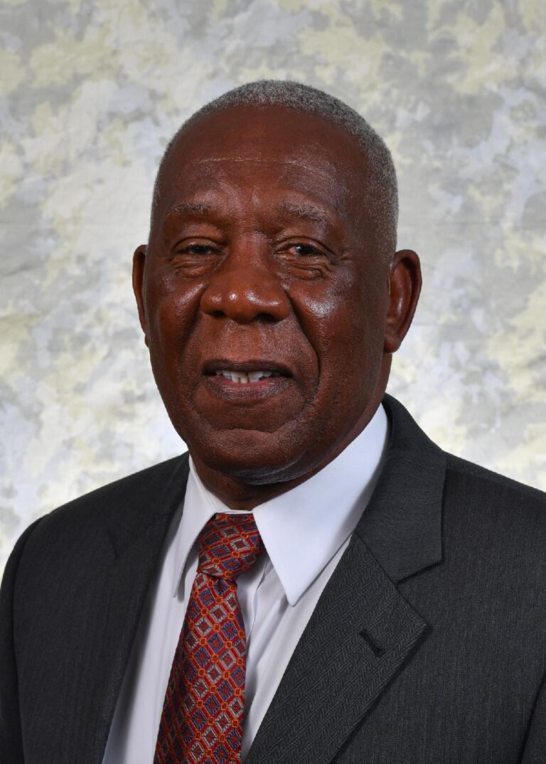 older man in a suit and tie poses for a professional portrait against a light, textured background.