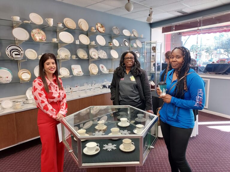 Three women stand around a glass display case with teacups in a store, with plates arranged on the wall in the background.