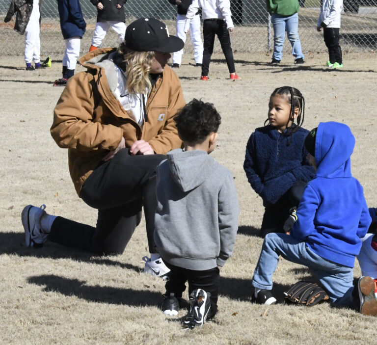 adult kneels and talks to three young children on a grassy field, with other people visible in the background, possibly during a sports practice or outdoor activity.