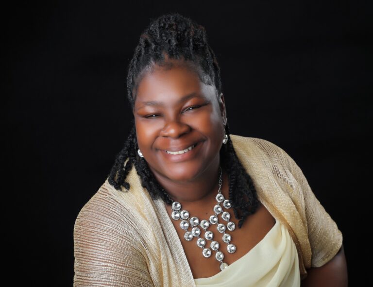 Smiling woman with braided hair wearing a light-colored dress, a gold shawl, and a statement necklace, posing against a black background.