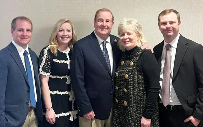 Four people stand close together indoors, smiling at the camera, dressed in formal and semi-formal attire against a plain light-colored background.