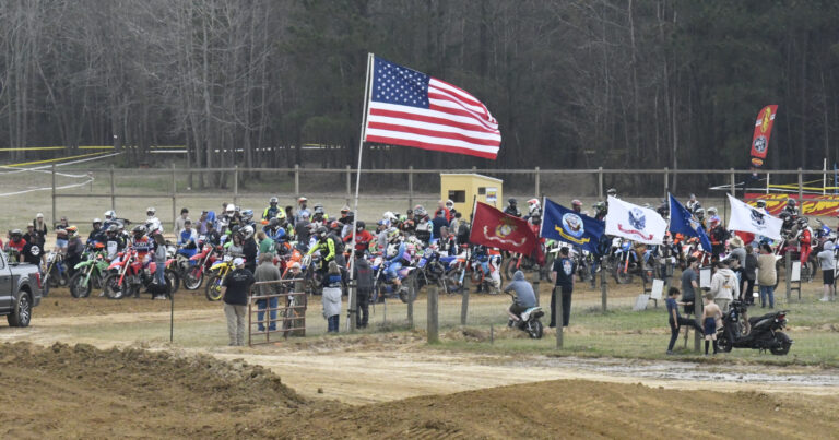 A group of motocross riders gather at a dirt track start line, with large American and military flags displayed, and forest in the background.