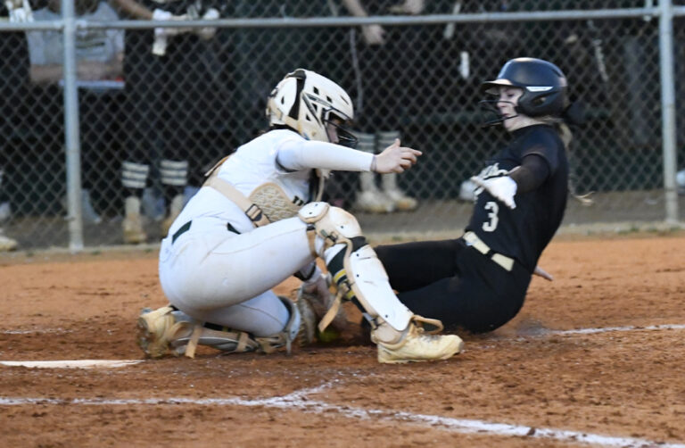 A softball catcher in white tags a sliding player in black at home plate during a game.