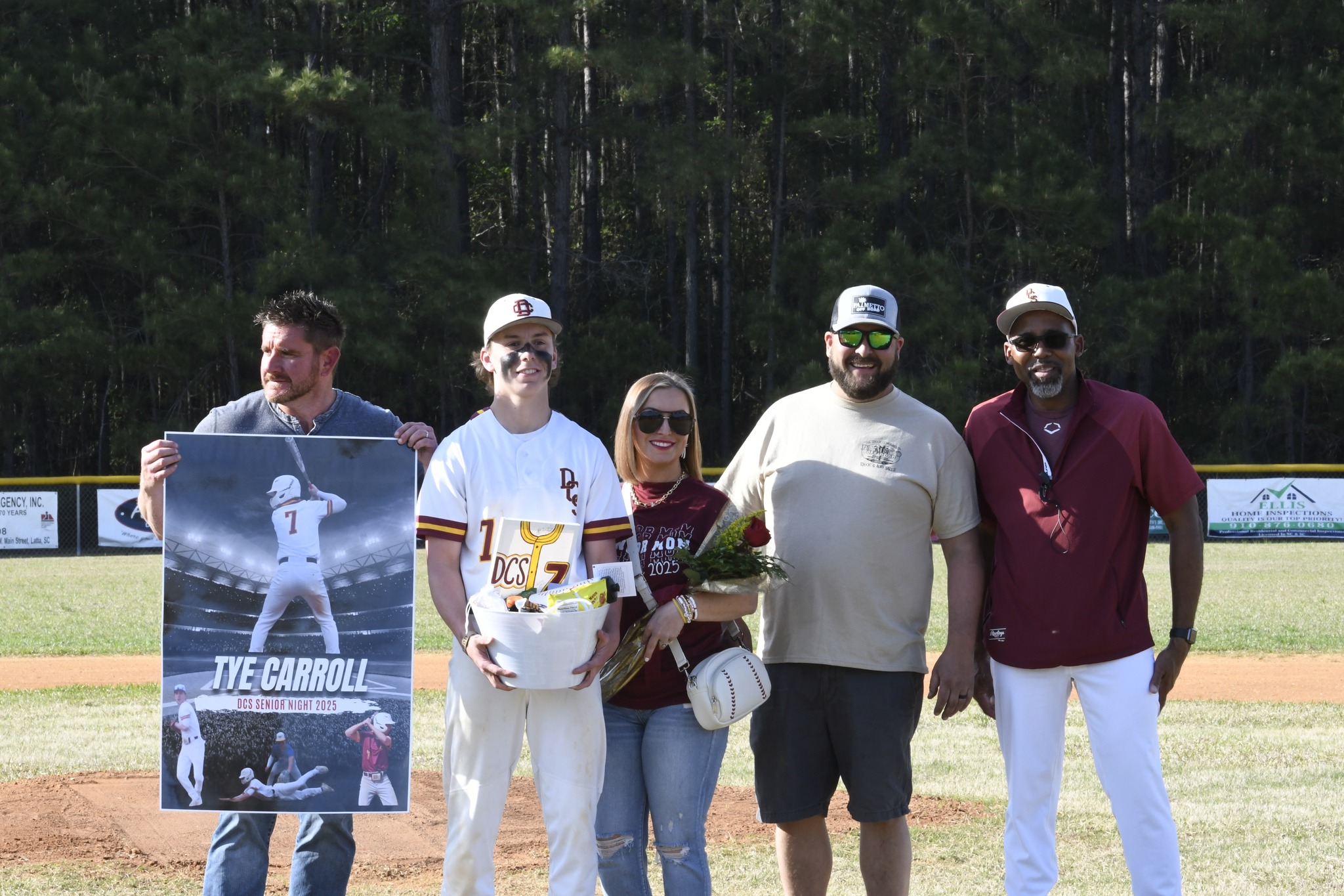 A group of six people stands on a baseball field, holding a poster, flowers, and a helmet, posing for a photo together.
