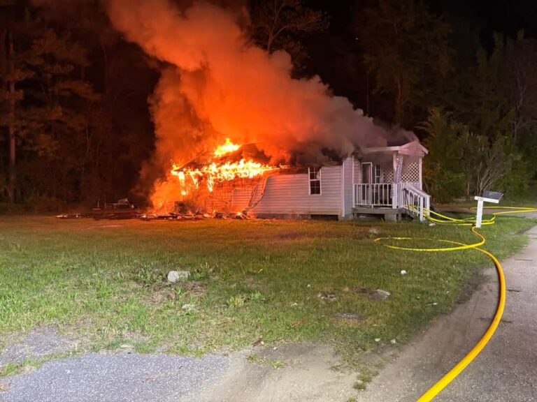 A house engulfed in flames at night with heavy smoke rising, surrounded by trees and a grassy yard.