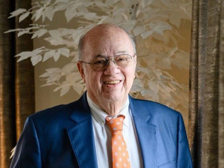 older man wearing glasses, a blue suit, and an orange polka-dot tie, smiling in front of a leafy patterned background.