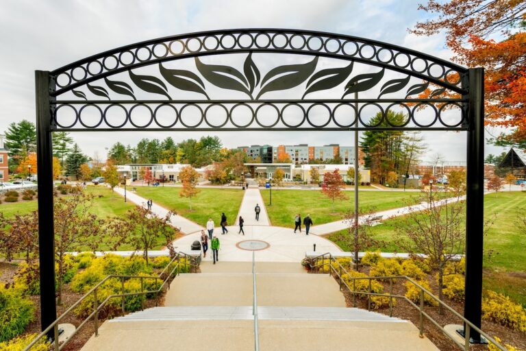 A college campus with a decorative arch, green lawns, walking paths, trees with autumn colors, and several people walking towards a modern building.