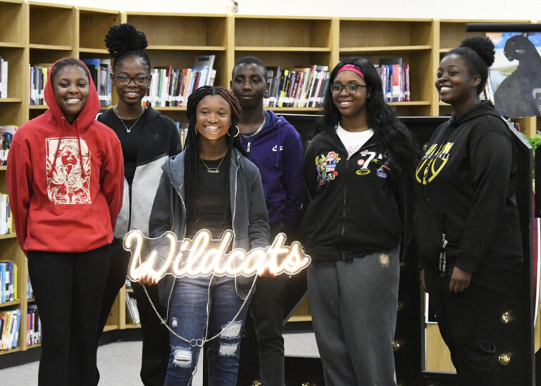 Six smiling students stand together in front of bookshelves. They wear hoodies and casual clothes. A neon sign in front reads “Wildcats.”