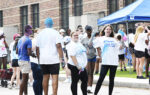 A group of people wearing white shirts and shorts gather outside near a brick building, some smiling and waving, at what appears to be a community event or race.