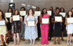 A group of women standing indoors, smiling and holding certificates, dressed in formal and semi-formal attire, posing for a graduation or award ceremony photo.