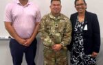Three people standing indoors: a man in a pink shirt, a person in military uniform, and a woman in a dark blazer, all smiling at the camera.