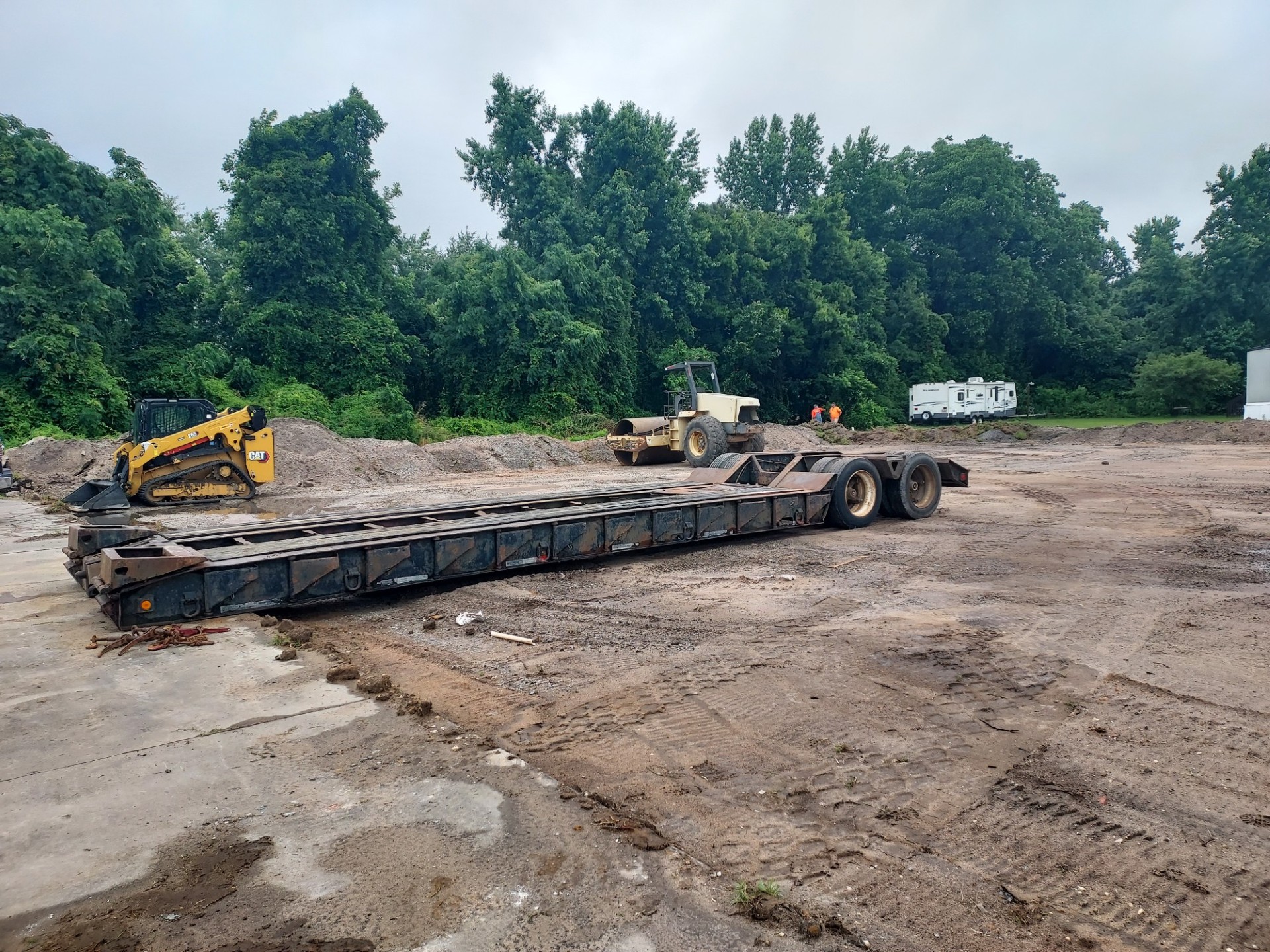 A large flatbed trailer parked on a dirt lot, surrounded by construction equipment and green trees in the background under a cloudy sky.