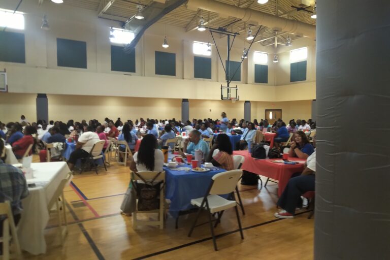 People seated at tables covered with red and blue tablecloths in a large gymnasium, attending an event or gathering.