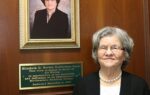 older woman stands in front of a wooden wall featuring her portrait photograph and a commemorative plaque.