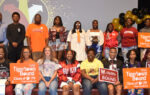 A group of students poses indoors, some holding college-bound signs and wearing college apparel, with balloons and a decorated backdrop behind them.