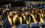 A group of graduates in caps and gowns, sitting and smiling at a graduation ceremony, some waving at the camera.