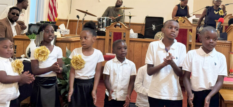 Three children stand in front of a church altar, wearing white tops, with a drummer and drum set visible in the background.