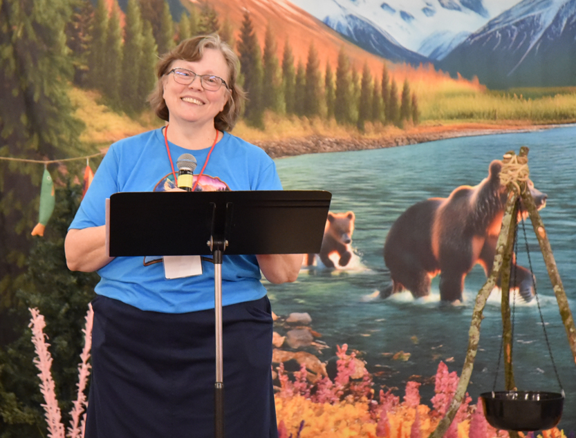 A woman standing at a podium, smiling, with a nature backdrop featuring bears, trees, a river, and mountains.