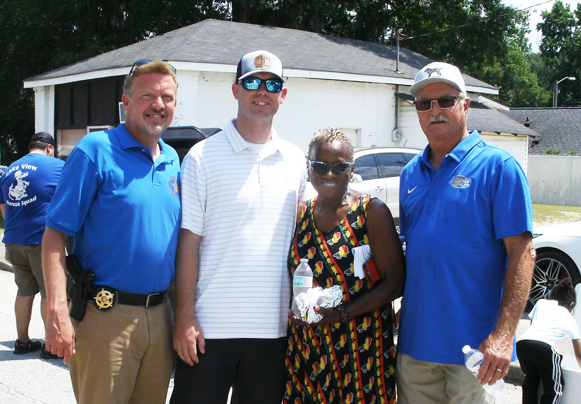 Four people stand together outdoors, three men in blue shirts and one woman in a patterned dress holding water bottles, smiling for the photo.