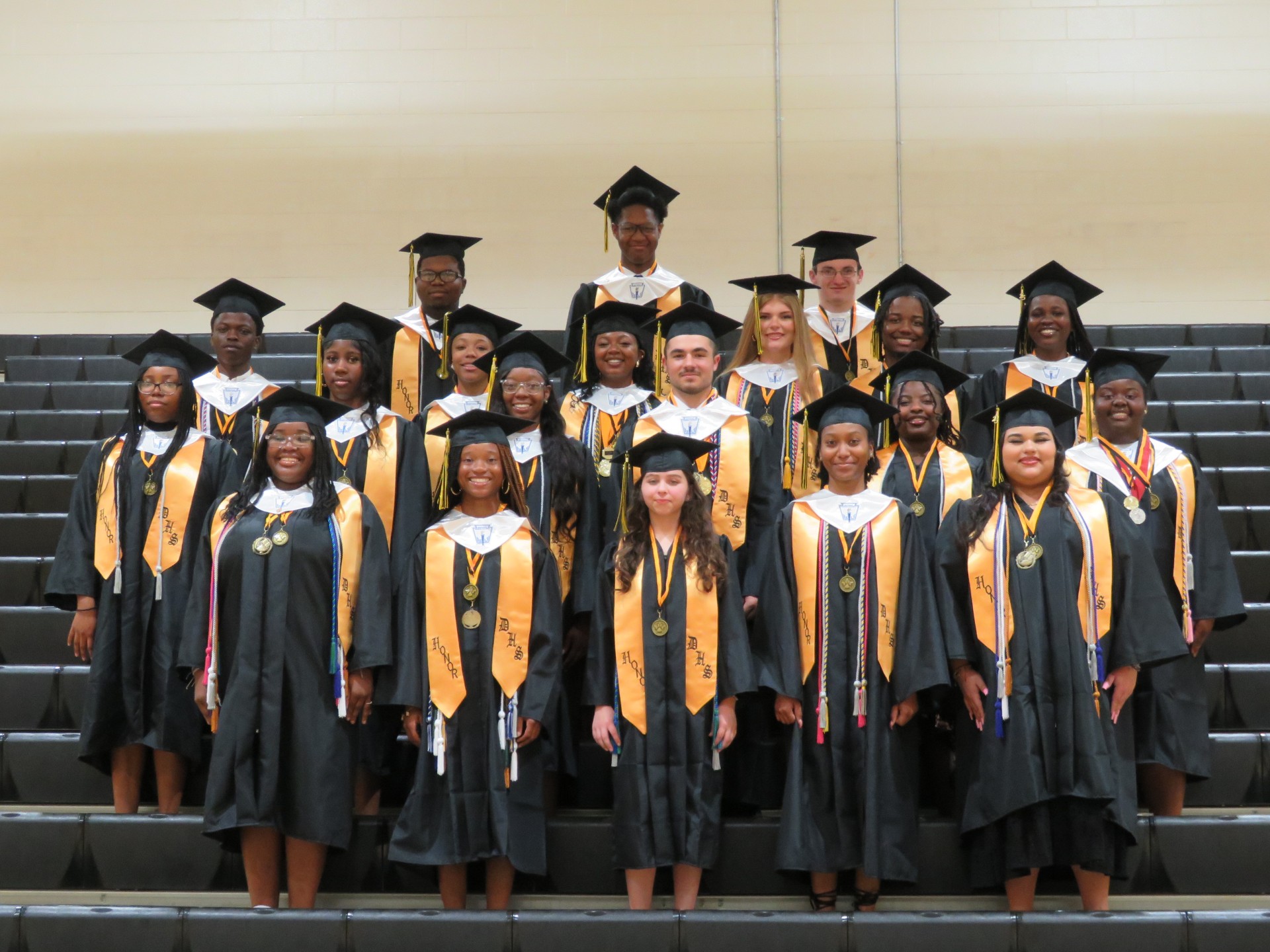 A group of graduating students in caps and gowns pose together, smiling for a class photo on a stage.