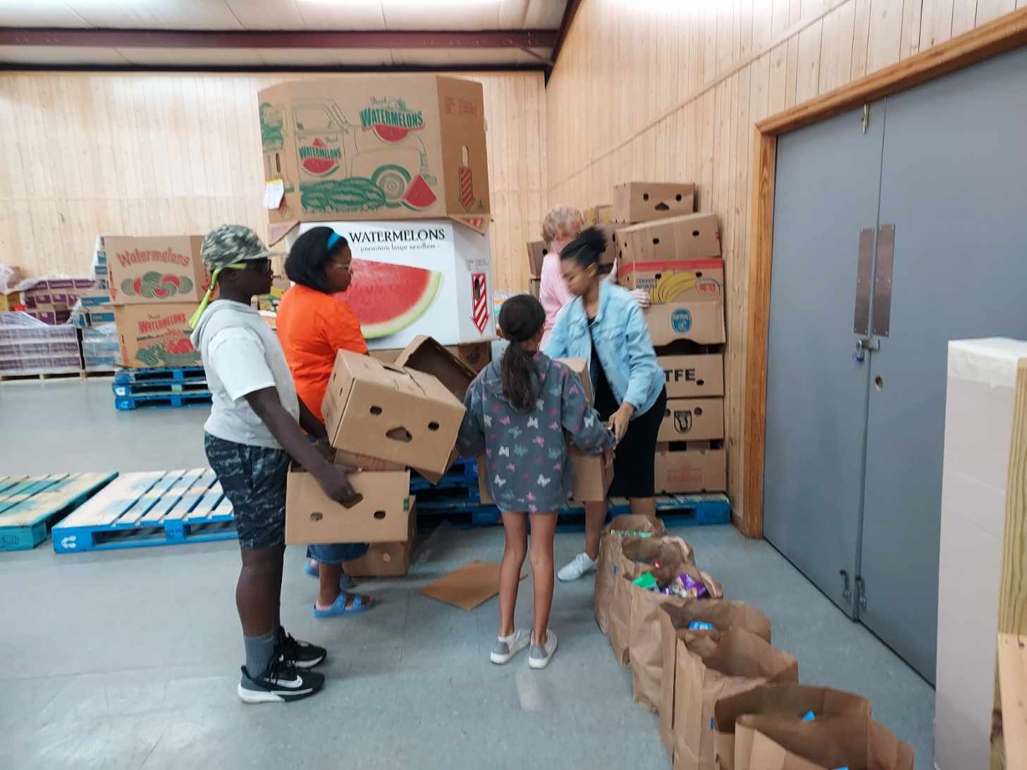Four people, including two children, organize and distribute boxes of food in a storage room lined with cardboard boxes and grocery bags.