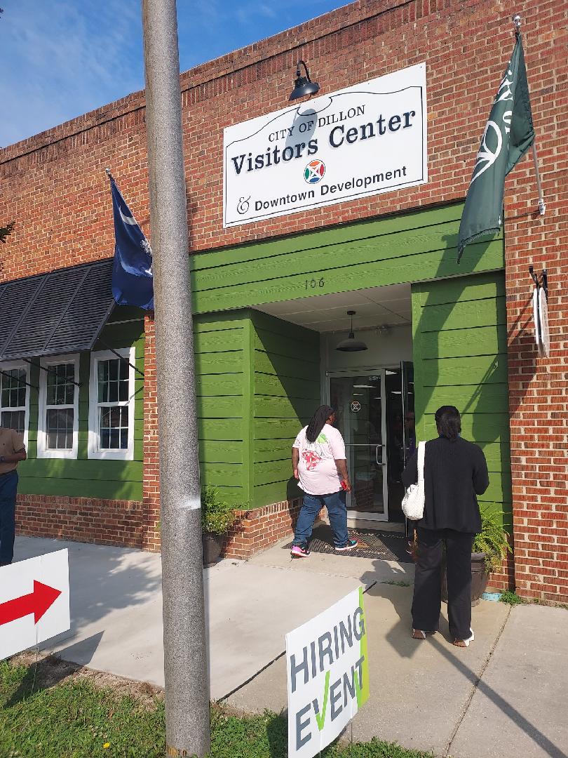 Two people enter a brick building with a green entrance labeled "Visitors Center." Flags hang outside, and a sign with an arrow points toward the entrance.