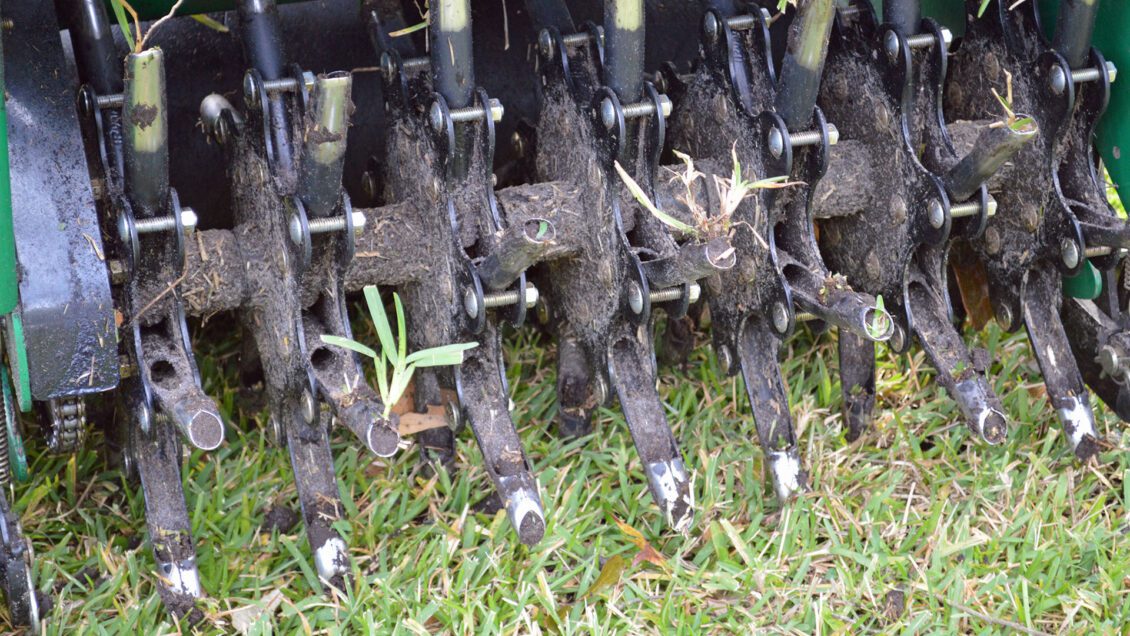 Close-up of a metal agricultural machine part with soil and grass stuck between its tines, resting on green grass.