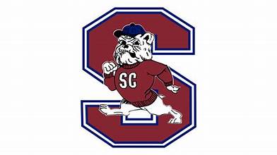 A bulldog wearing a red sweater with "SC" and a blue cap, standing in front of a large red letter "S" outlined in blue and white.