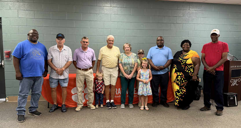 A group of adults and children stand in a line indoors, posing for a group photo against a green wall with an orange tablecloth in the background.