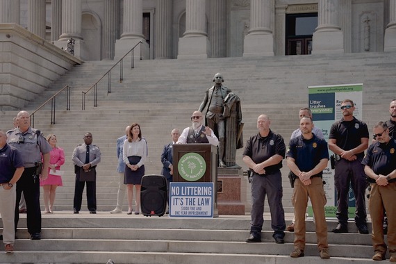 A group of people stands on government building steps during a press conference, with a statue and signs visible in the background.