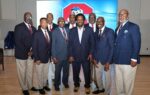 Nine men in suits and ties pose together indoors, smiling, with a red and blue logo displayed on a screen behind them.
