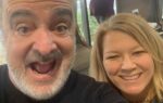 A man with a surprised expression and a woman smiling pose for a selfie indoors, with blurred people and windows in the background.