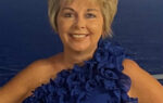 Smiling woman wearing a blue ruffled one-shoulder dress stands in front of a blue ocean background.