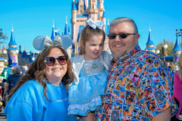 A family of three smiling in front of a castle at a theme park, with the adults wearing sunglasses and the child dressed as a princess.