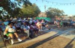 A group of people sitting outdoors on benches in a park, enjoying a sunny day with string lights overhead and trees in the background.