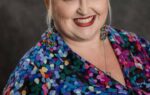 Smiling woman with blonde hair in a bun, wearing a colorful abstract-pattern blouse and earrings, poses in front of a dark studio background.