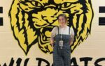 A person stands in front of a large yellow and black wildcat mural painted on a wall, with "WILDCATS" written below.