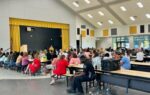 A large group of people seated at tables in a school cafeteria, listening to a speaker on stage with a yellow curtain backdrop.