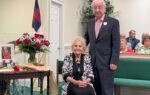 elderly woman sits beside an elderly man standing in a church, with flowers and a flag in the background. People are seated behind them.