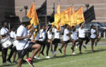 A group of people in white shirts and black shorts practice flag routines outdoors, holding gold and black flags, marching on a grassy field near a building.