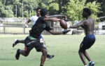 Three young men play football outside on a grassy field, one trying to catch a pass while another attempts to block him.