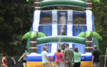 People gather in front of a large inflatable water slide decorated with palm trees, outdoors on a sunny day.