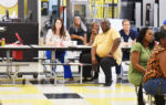 A group of adults sitting at cafeteria tables, attentively listening to someone out of frame, in a brightly lit room with yellow and black tiled floor.