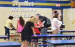 People gather in a school cafeteria, talking and filling out papers. Blue tables and a wall with the words “Together We Can Do Great Things” are visible.