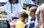 A woman hands out school supplies to two children outdoors, while an adult and another child watch nearby.
