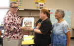 Three women standing in a classroom, one woman hands a framed photograph to another while the third woman looks on and smiles.