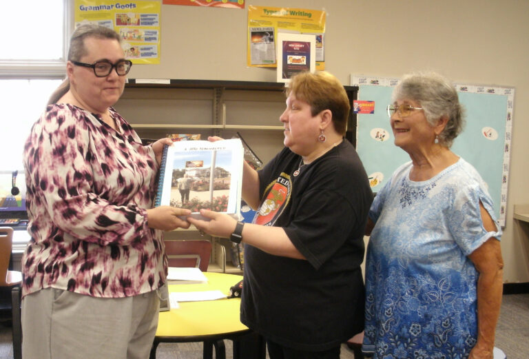 Three women standing in a classroom, one woman hands a framed photograph to another while the third woman looks on and smiles.