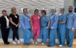 A group of seven healthcare workers in scrubs and uniforms stand in a line, smiling inside a medical facility.