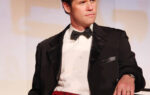 A man wearing a black tuxedo and red cummerbund leans on a chair while standing on a stage.